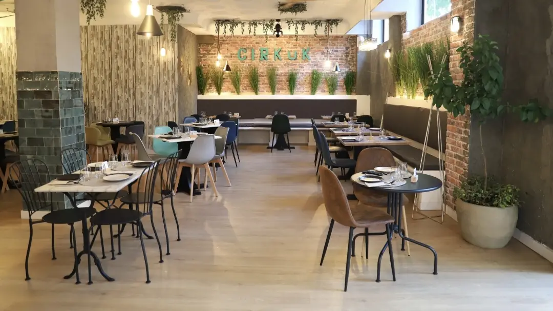 Restaurant dining room with wooden tables, upholstered chairs, and hanging pendant lights over a green wall.