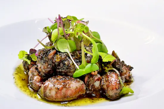Braised octopus pieces with herb oil and microgreens on a white plate, Clandestino