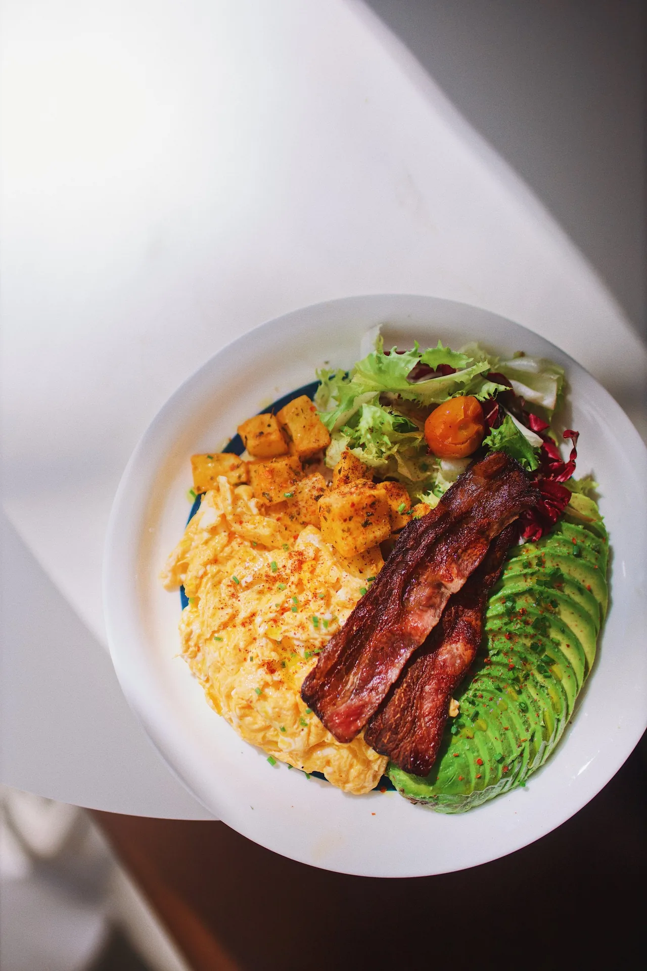 A top-down bowl of scrambled eggs, bacon, avocado and salad leaves
