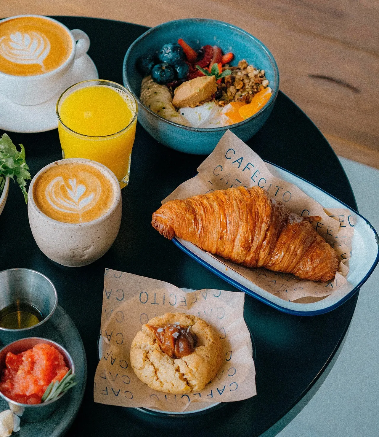 A breakfast table with a croissant, coffee, orange juice, a granola bowl and a cookie