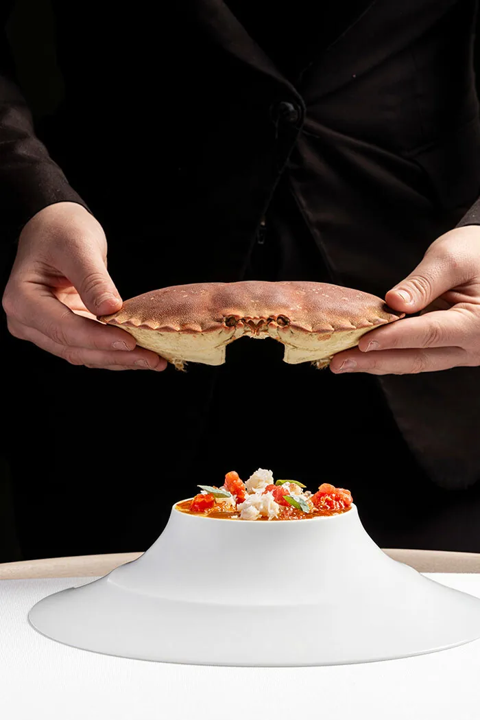 Cook in black holding an empty crab carapace above a plated crab dish with diced tomato and basil on a white conical stand.