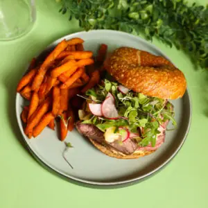 Steak sandwich with greens and radish on a brioche bun beside sweet potato fries on a grey plate