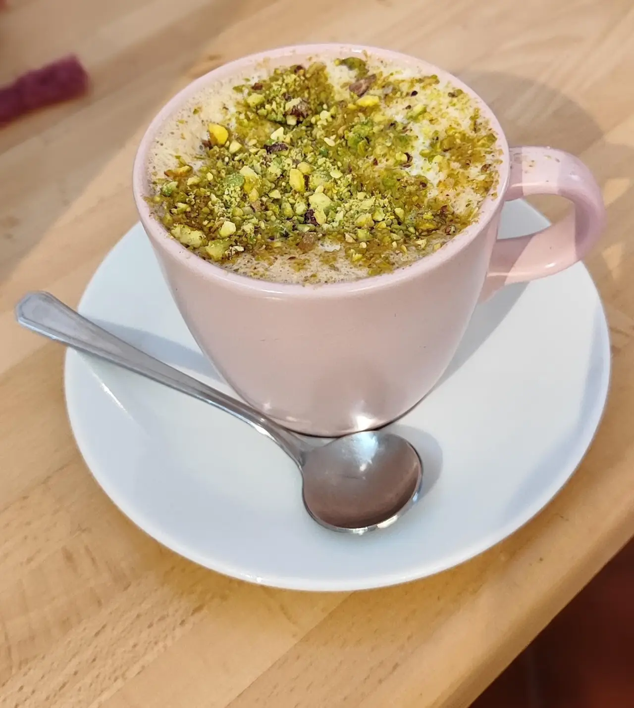 A latte topped with chopped nuts in a pink cup with a spoon on the saucer