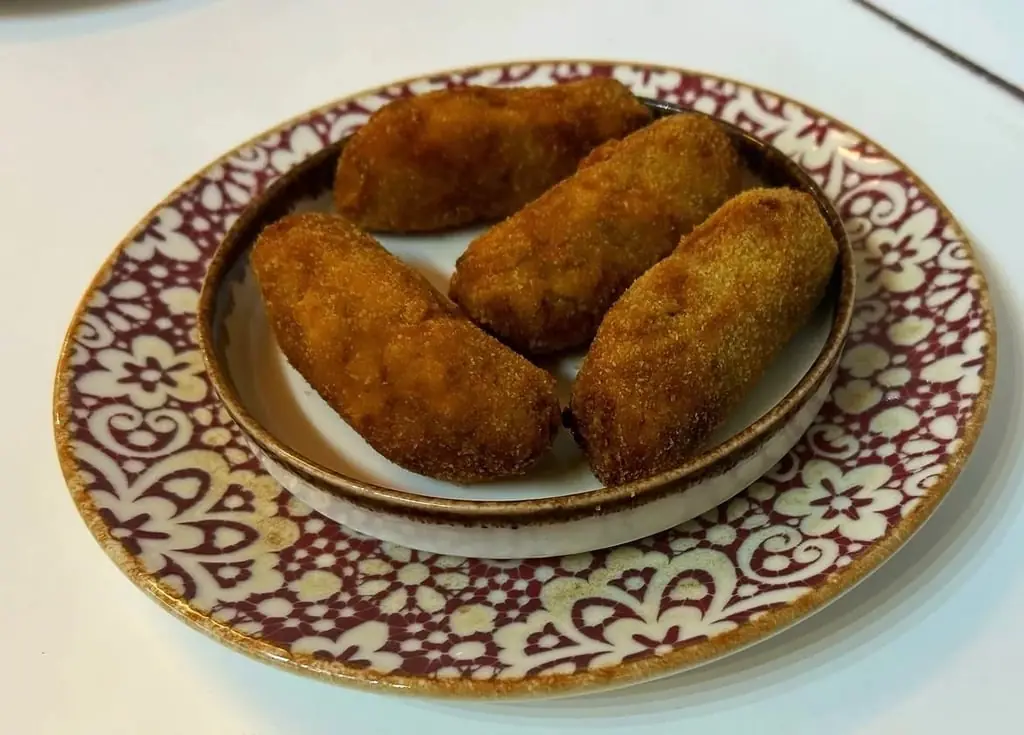 Four breaded and fried croquetas served in a small bowl on a patterned plate, Colmado Carpanta
