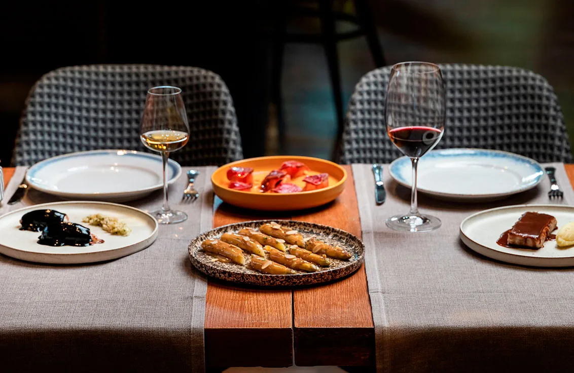 A table laid for two with a linen runner and a raw wooden centre plank, set with a glass of white and a glass of red wine, a shallow orange bowl of sliced red peppers, a speckled plate of pale vegetable batons in sauce, a small plate of dark puree with a herbed garnish and a glazed pork belly with a puree quenelle