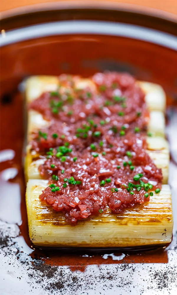 A charred split leek in a dark glossy reduction in an earthenware dish, blanketed with chopped raw red meat, chives and flakes of salt