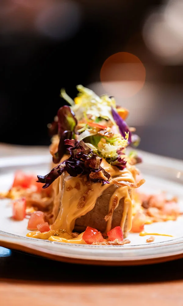 A tall fried block glazed with orange sauce, crowned with a tangle of mixed leaves and crisp shreds, ringed by diced tomato on a pale ceramic plate