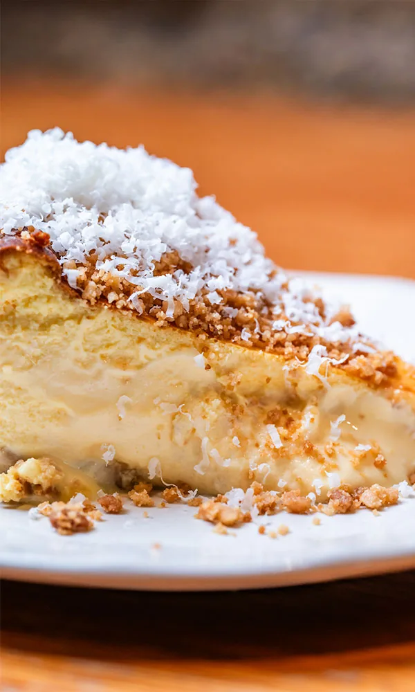 A wedge of soft-set baked cheesecake with a molten centre, its top crusted with crumb and blanketed in finely grated white cheese, on a white plate on a wooden table