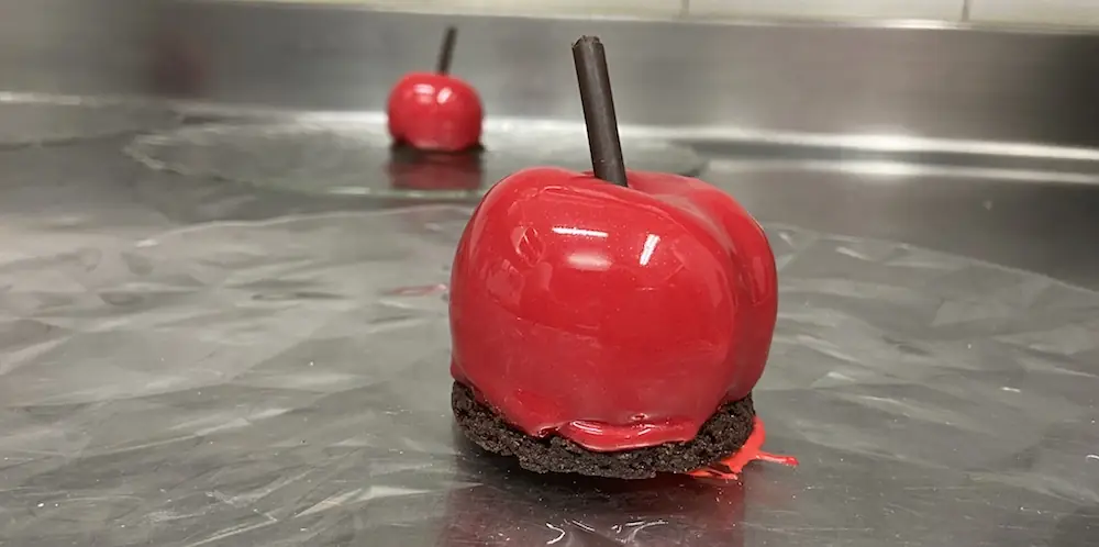 Glossy red candy-apple dessert with a chocolate stick, on a chocolate-crumb base, Con Gracia