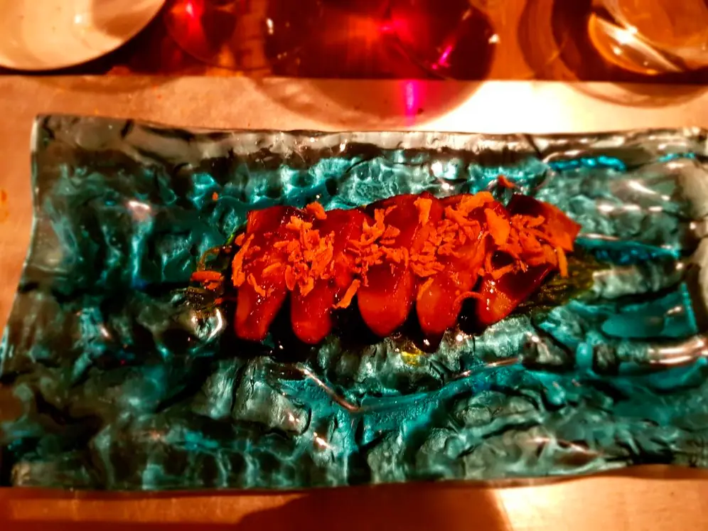 Sliced glazed dish topped with crispy shredded garnish, plated on a textured slate tray, Con Gracia