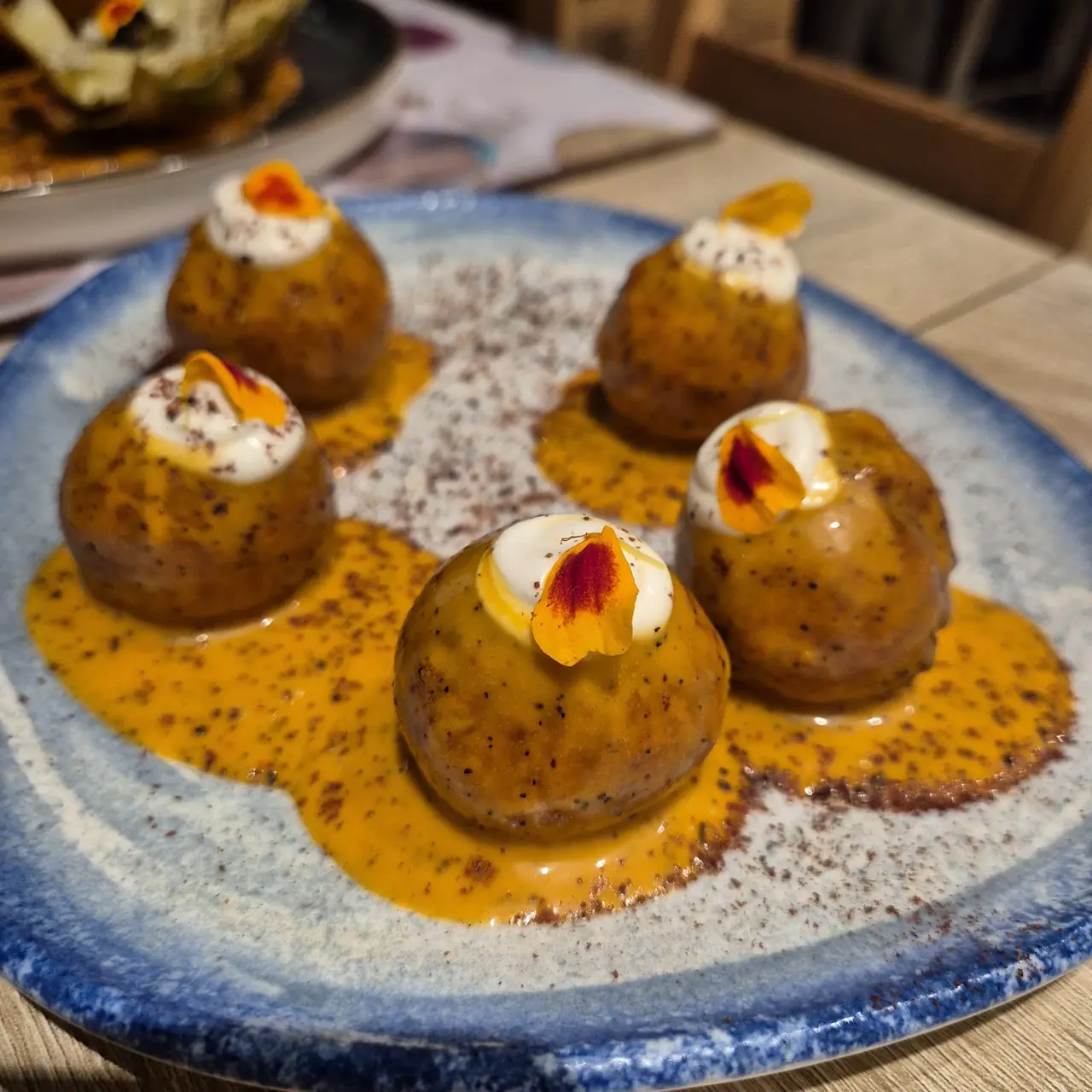Five roasted potato-like croquettes topped with cream and edible flower petals in orange sauce on a blue plate