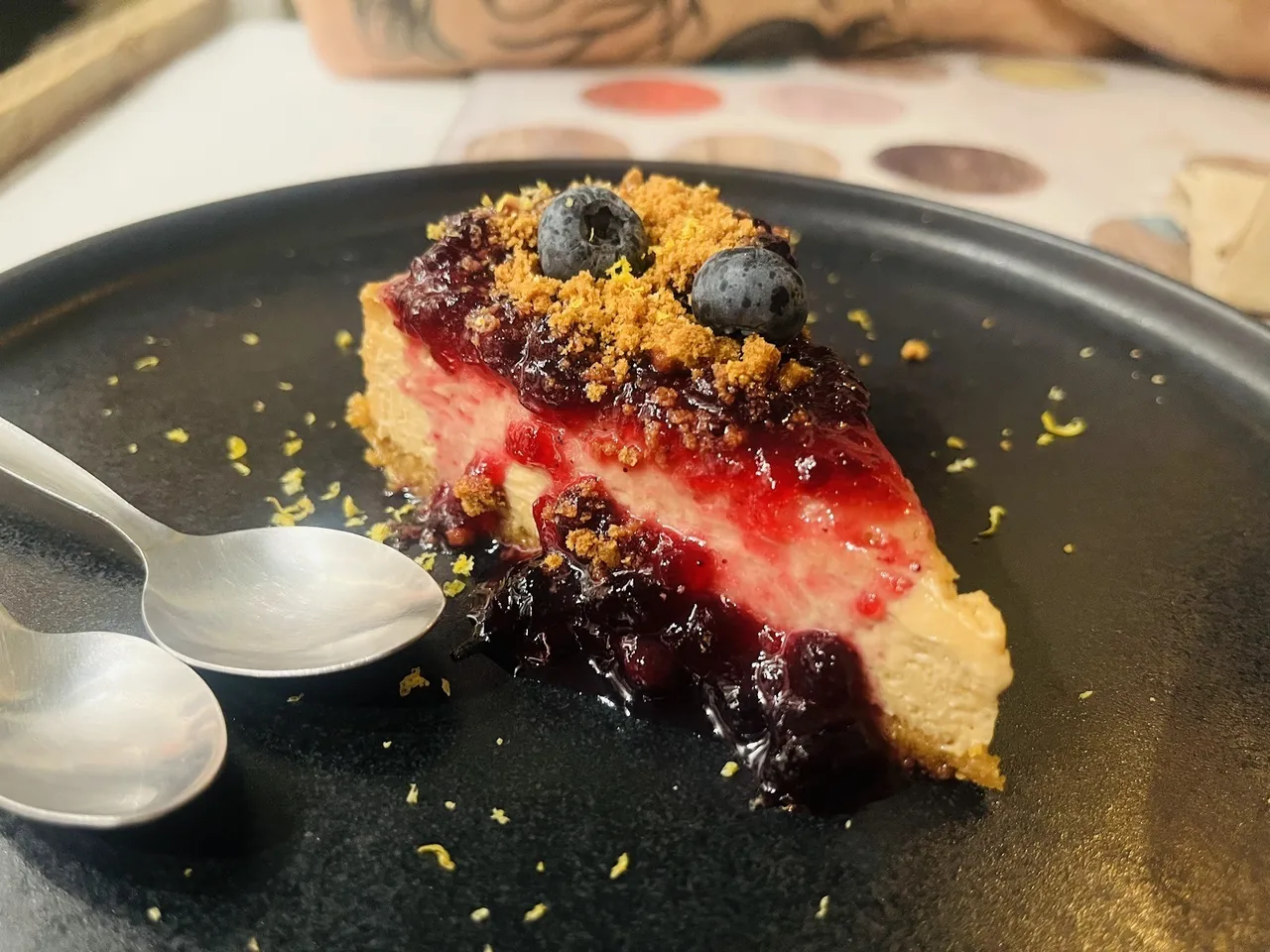 A slice of cake with a dark berry topping and crumbs on a dark plate