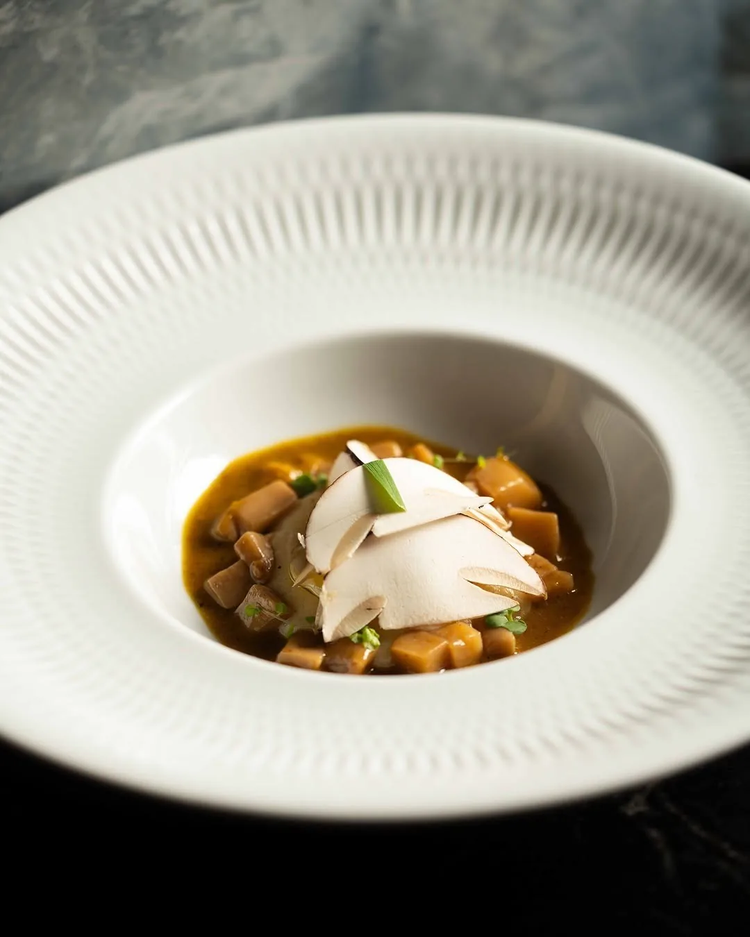 A white ribbed bowl holding a broth with diced vegetables and shaved slices on top
