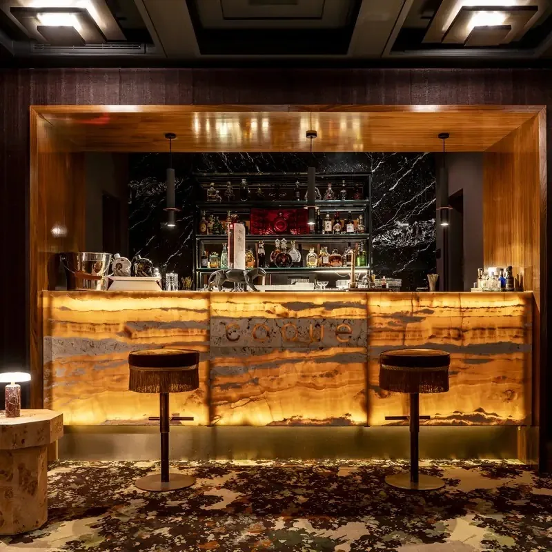 A bar with a glowing amber front, two stools and shelves of bottles behind it