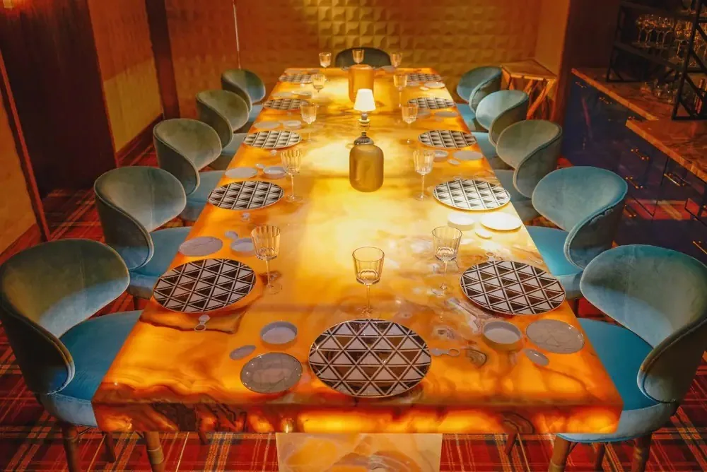 A long table with an amber glowing top, blue velvet chairs and patterned plates