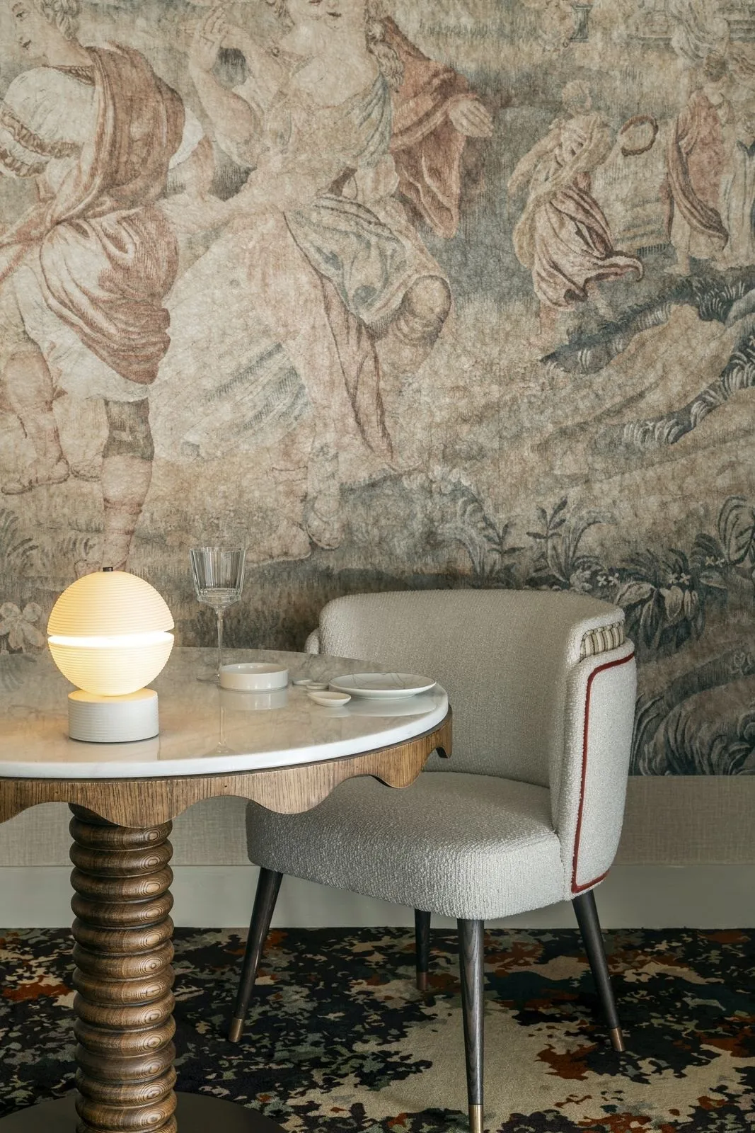 A small round table with a glowing lamp in front of a cream armchair and an ornate patterned wall