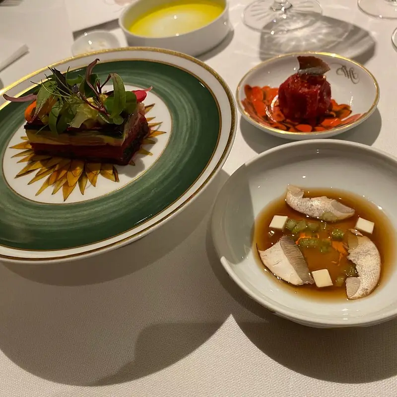Three plated dishes on table: layered vegetable terrine, small braised dish, and broth with fish and vegetables
