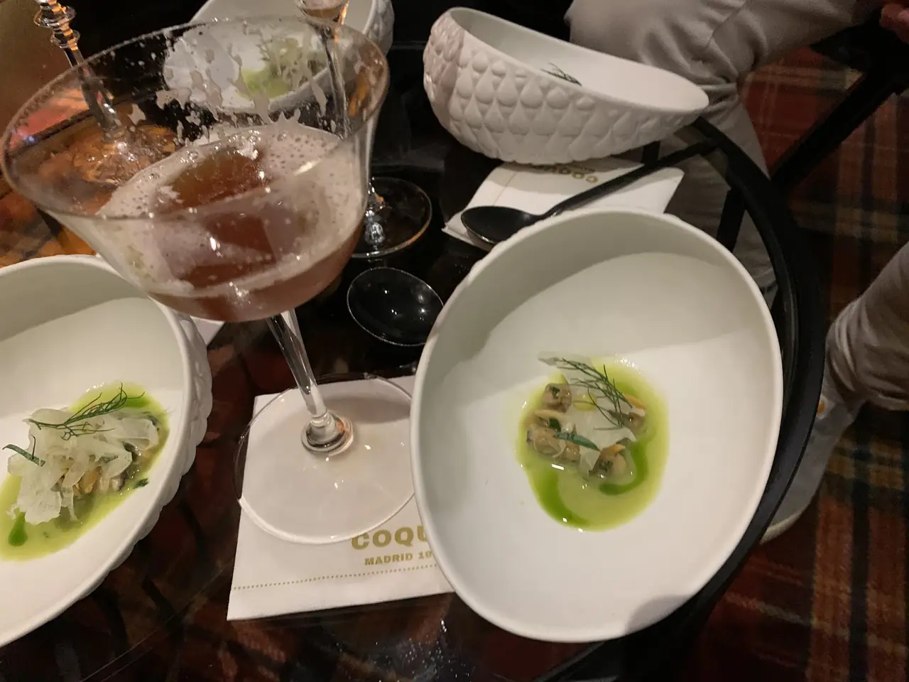 Small plated dishes with green sauce and shellfish beside a poured cocktail in coupe glass on table