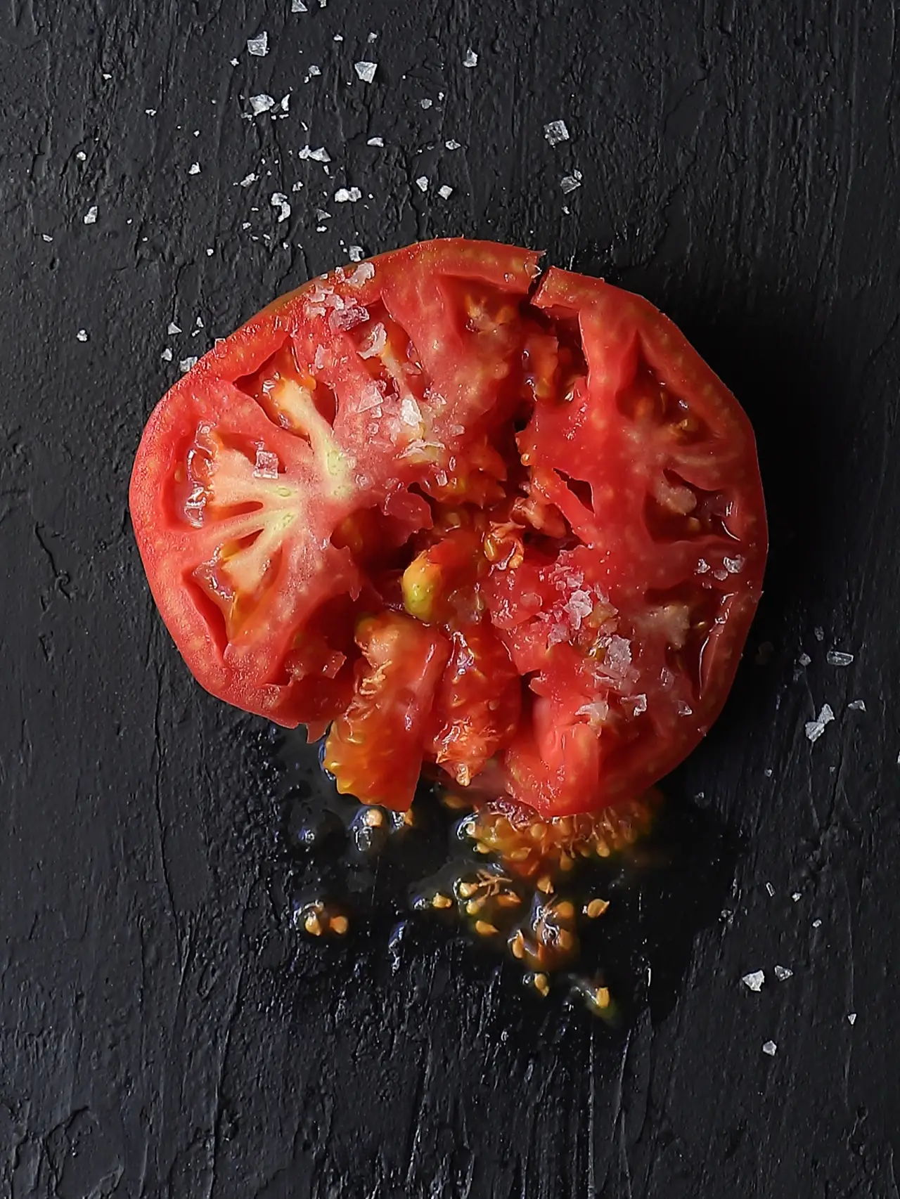 A halved heirloom tomato scattered with flaky sea salt on a black slate surface