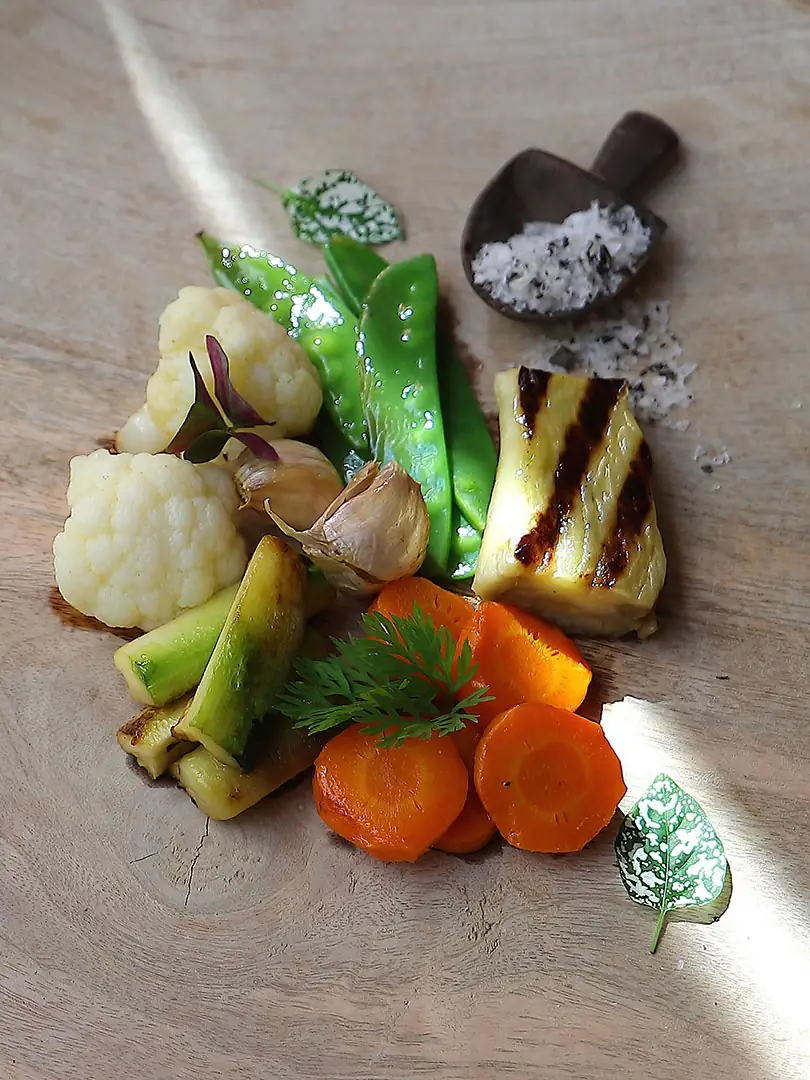 Plated grilled seasonal vegetables including cauliflower, snap peas, carrots, garlic and courgette with a scoop of dark salt on a wooden board
