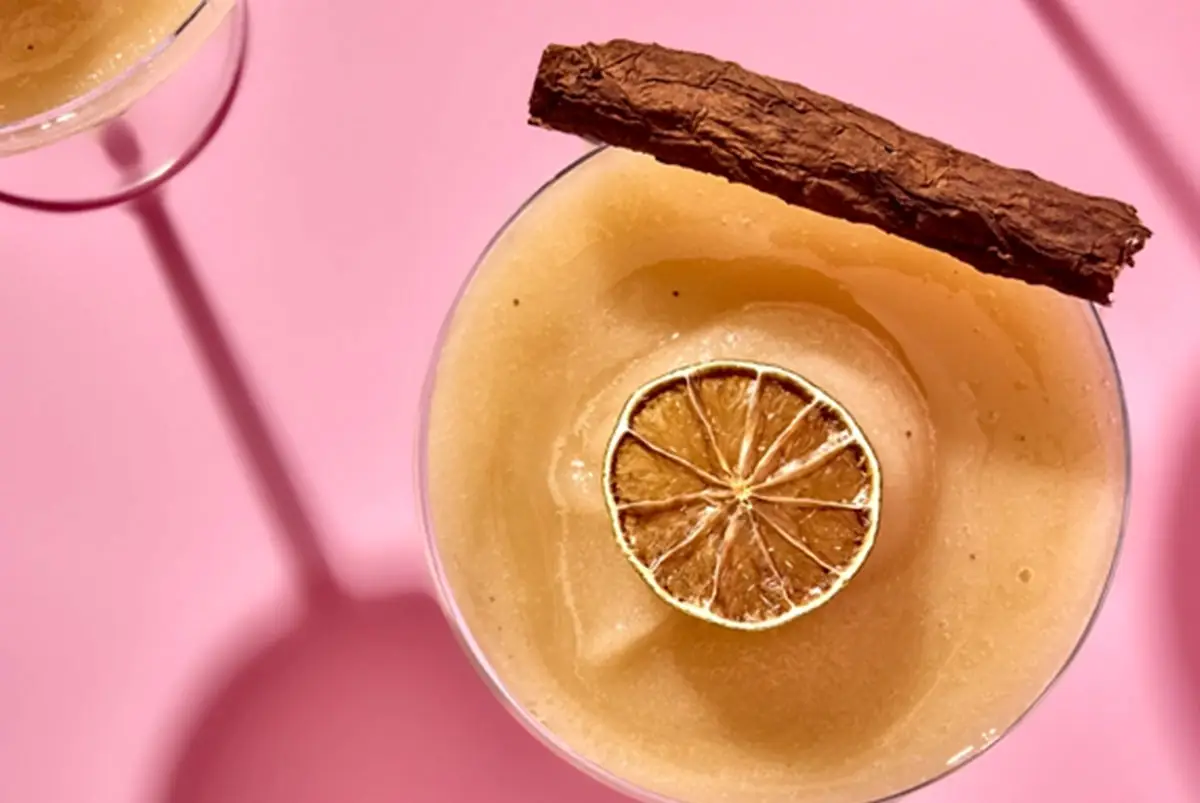 Overhead of a frothy amber cocktail in a coupe garnished with a dried citrus wheel and a cinnamon stick on a pink backdrop