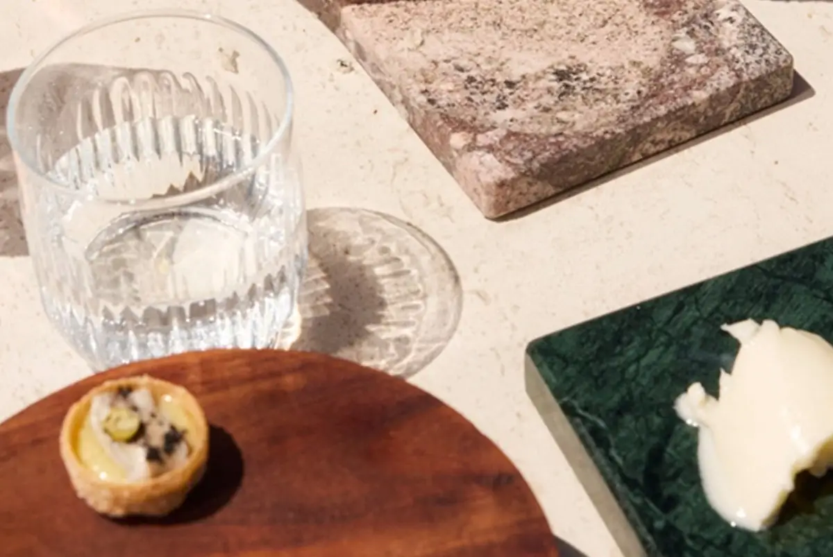 Sunlit table with a small tartlet on a wooden board, a cut-crystal water glass and a quenelle of butter on a green marble slab