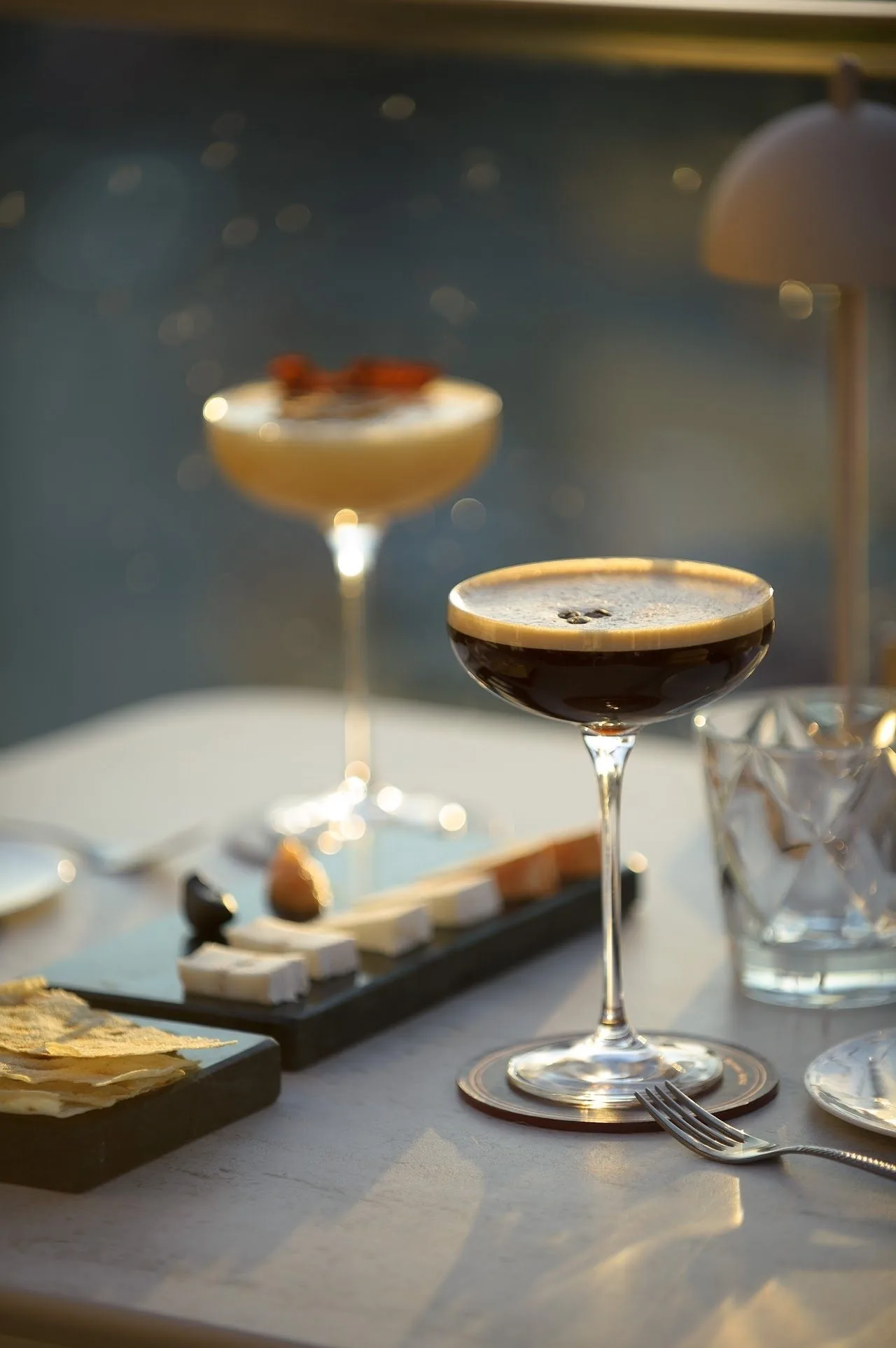 An espresso martini in a coupe glass on a table beside a small plate of sweets