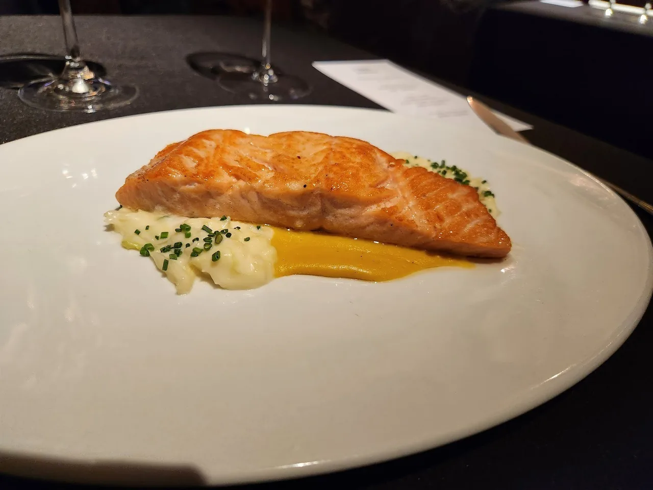 A piece of pan-seared orange fish on a white plate with a yellow sauce and chives