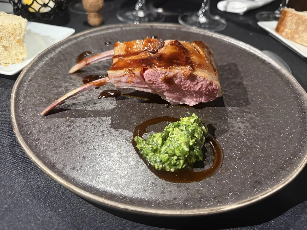 A rack of lamb ribs on a dark plate beside a green herb sauce