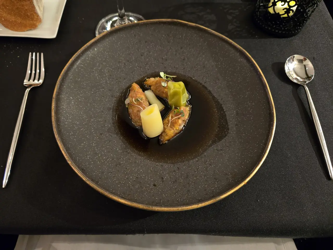 Braised mussels with a dark reduction sauce and leek batons in a wide dark bowl on a formal table setting