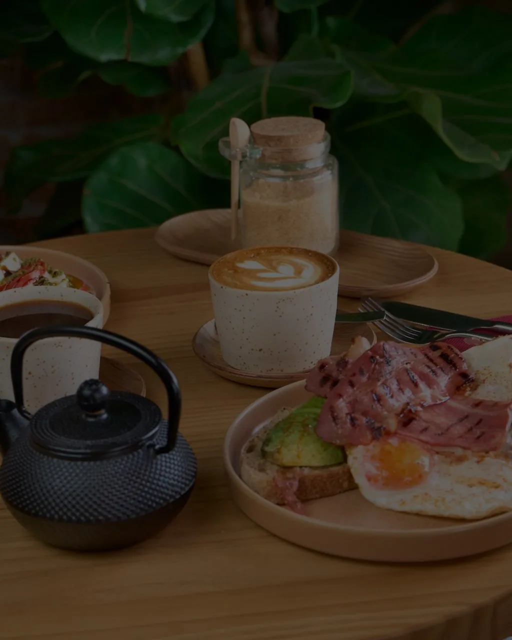 A breakfast table with a latte, a plate of toast with egg and bacon and a black teapot