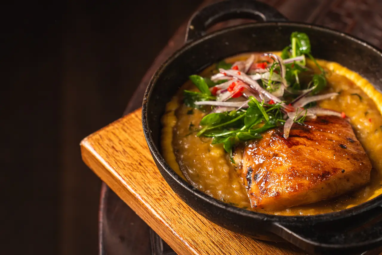 Seared fish fillet with grilled char marks on a corn and bean stew, topped with pickled red onion, chili strips, and pea shoots in a cast-iron pan, Coya