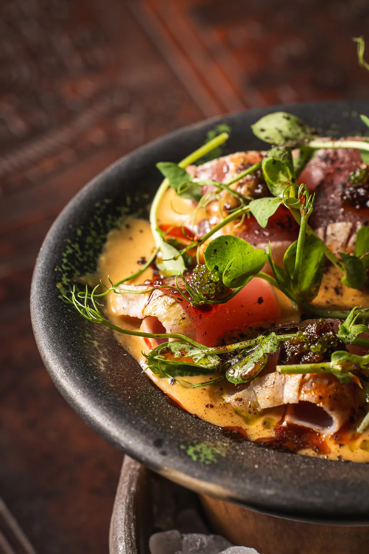 Seared tuna slices with charred edges over a creamy orange sauce, garnished with tomato, black tobiko, herb oil, and microgreens on a dark ceramic plate, Coya