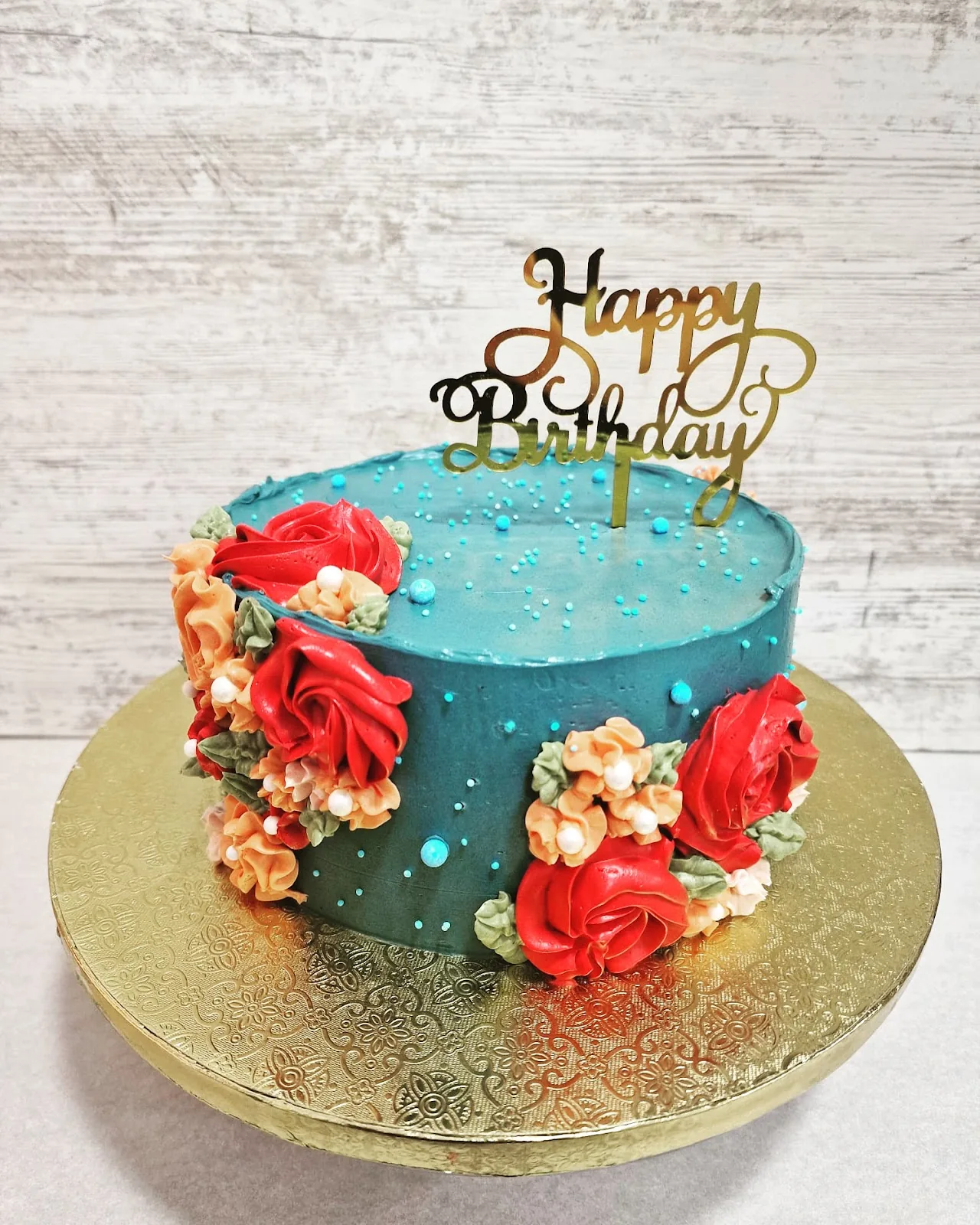 A round birthday cake decorated with red sugar flowers and a gold cake topper