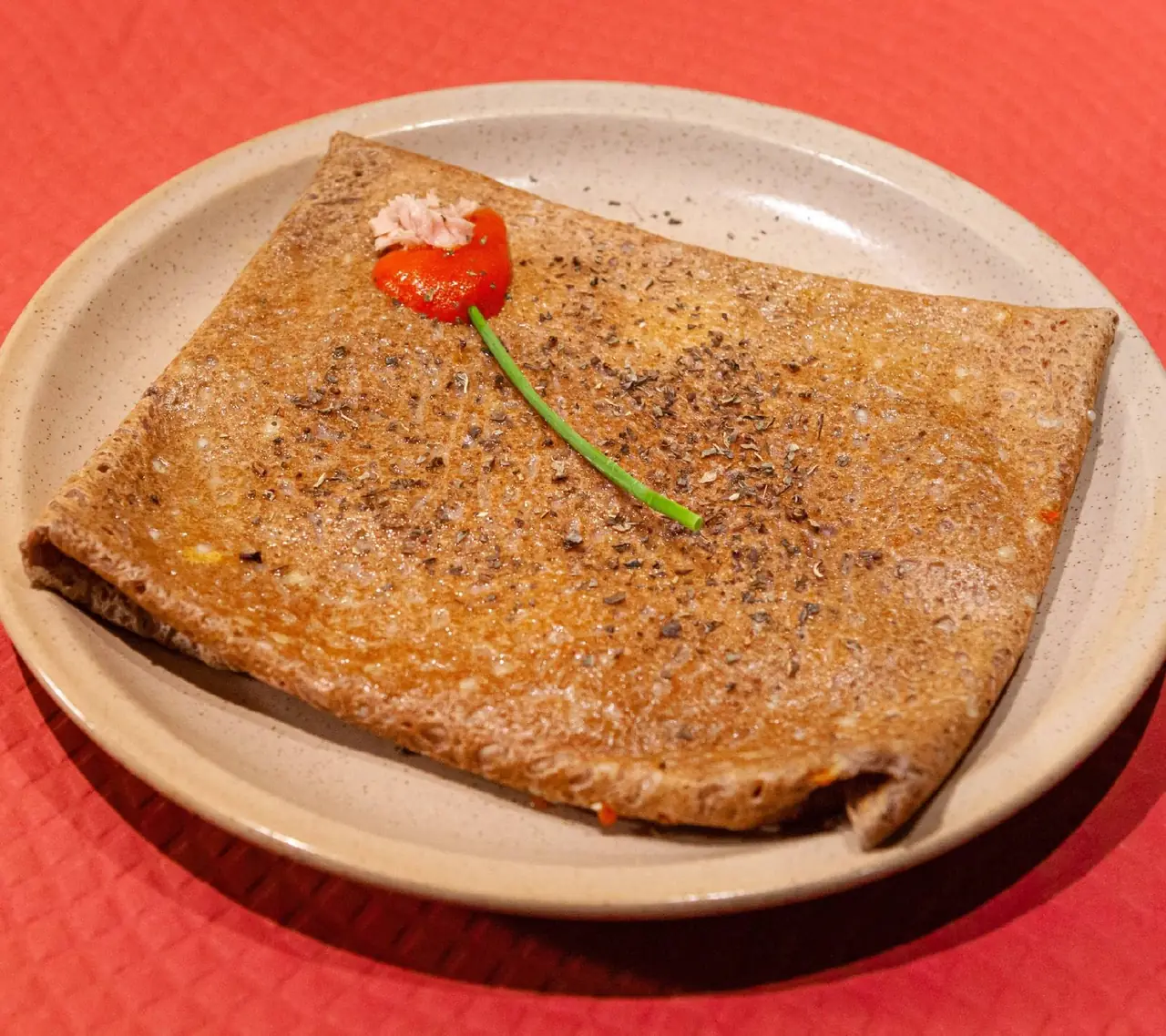 Folded buckwheat galette topped with ham, a slice of red pepper, and chopped chives on a stoneware plate, at Crêperie Les 3 Pommes