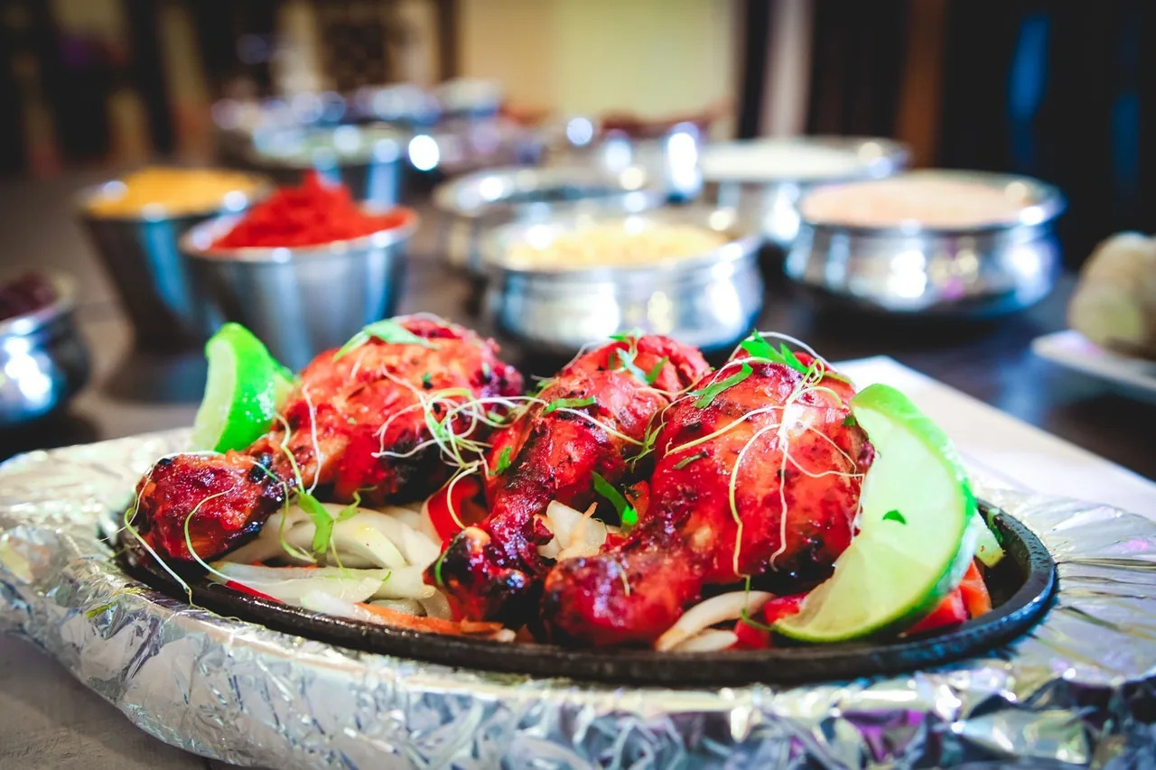 Red grilled chicken drumsticks with lime wedges on a sizzling plate lined with foil, with metal bowls of spices behind