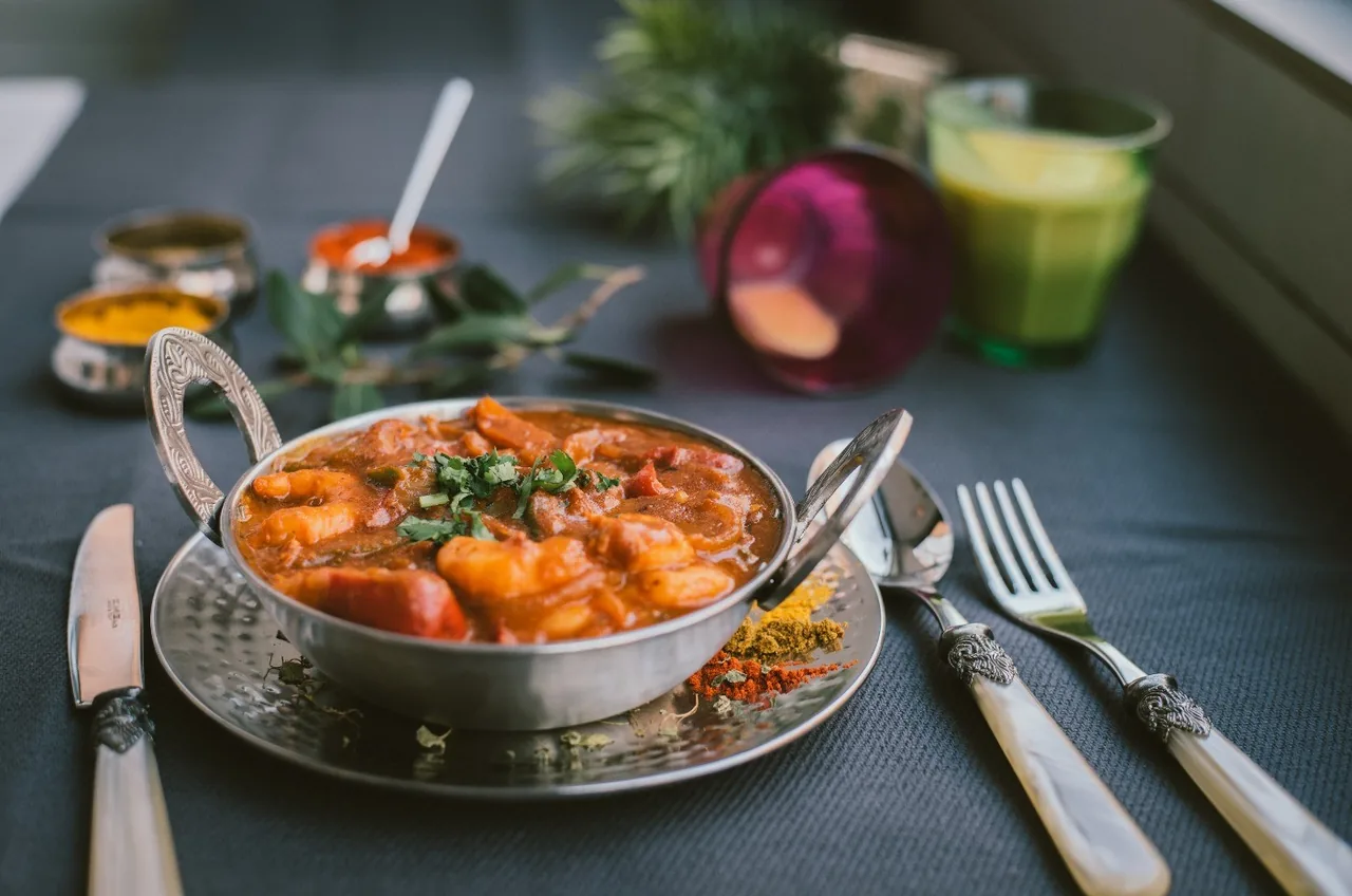 A metal karahi bowl of orange curry with prawns and coriander on a hammered plate, with spices and cutlery on a grey cloth