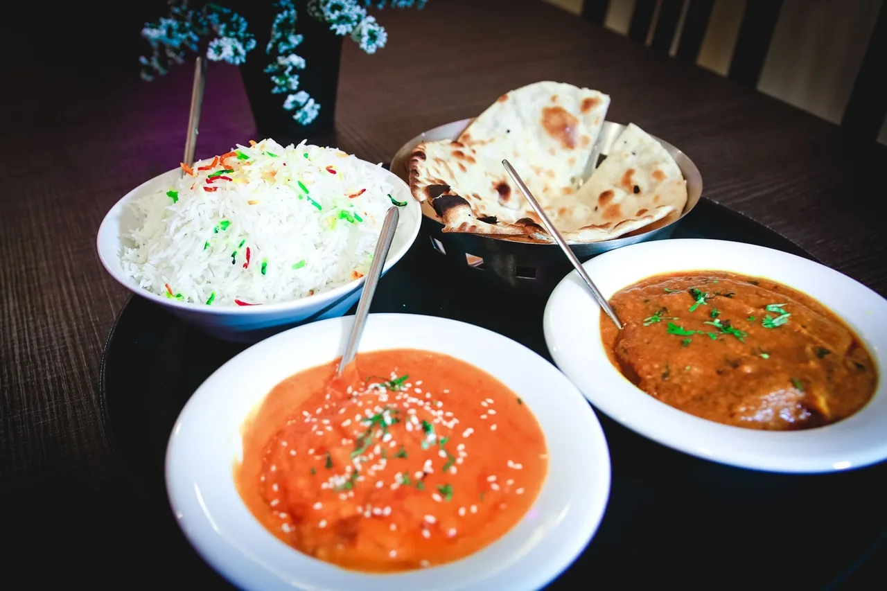Two white bowls of orange and brown curry beside a bowl of rice and a basket of naan on a dark table