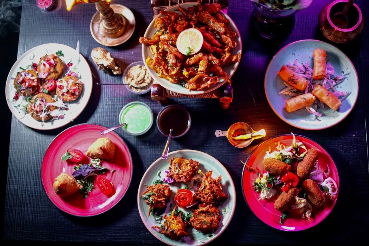 A dark table seen from above with several plates of fried snacks, samosas, fritters and small bowls of chutney