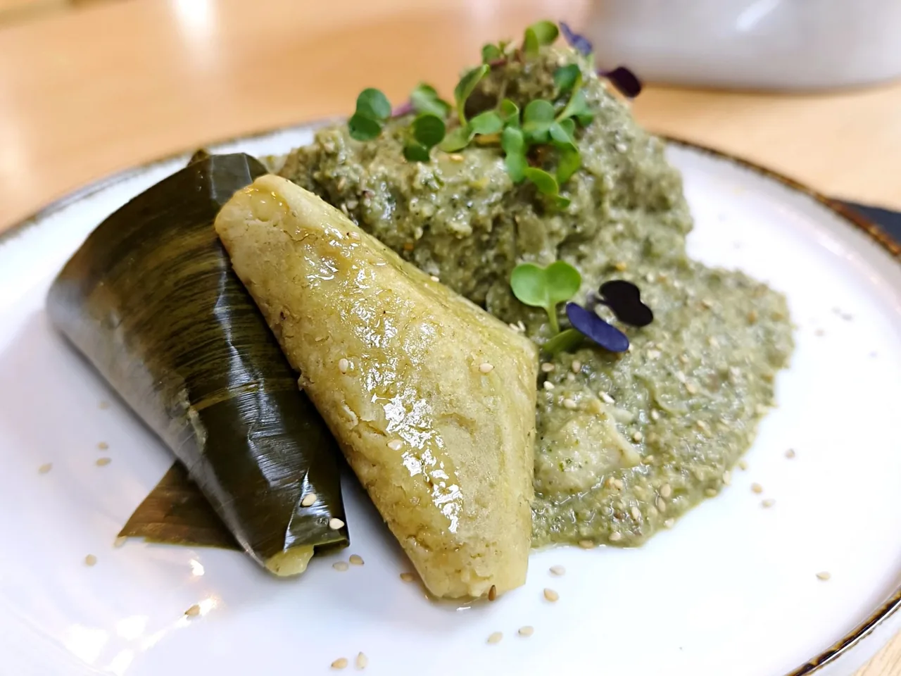 A leaf-wrapped tamal on a white plate served with a green sauce and micro herbs