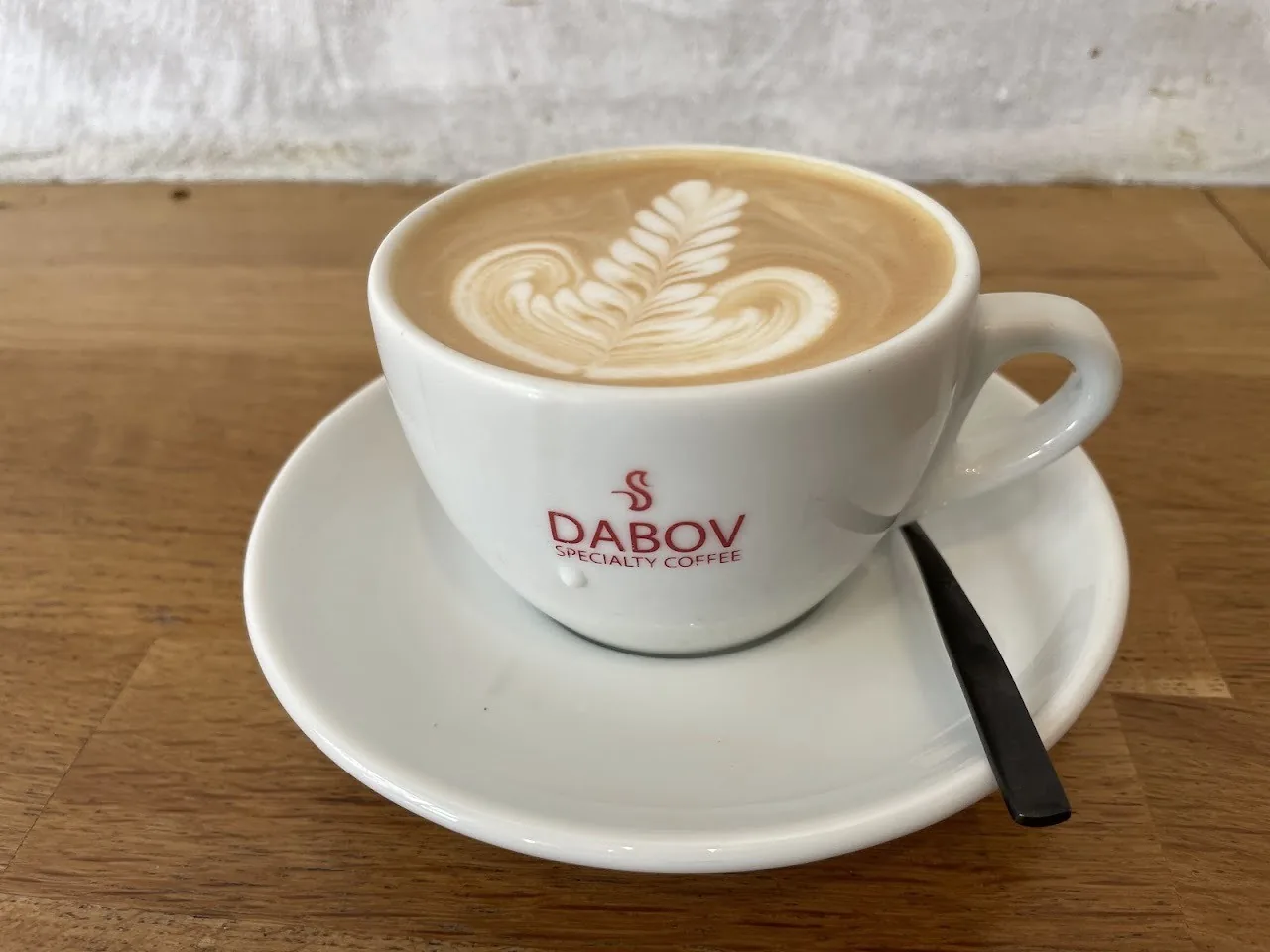 A latte with a leaf pattern in a white branded cup and saucer with a black spoon