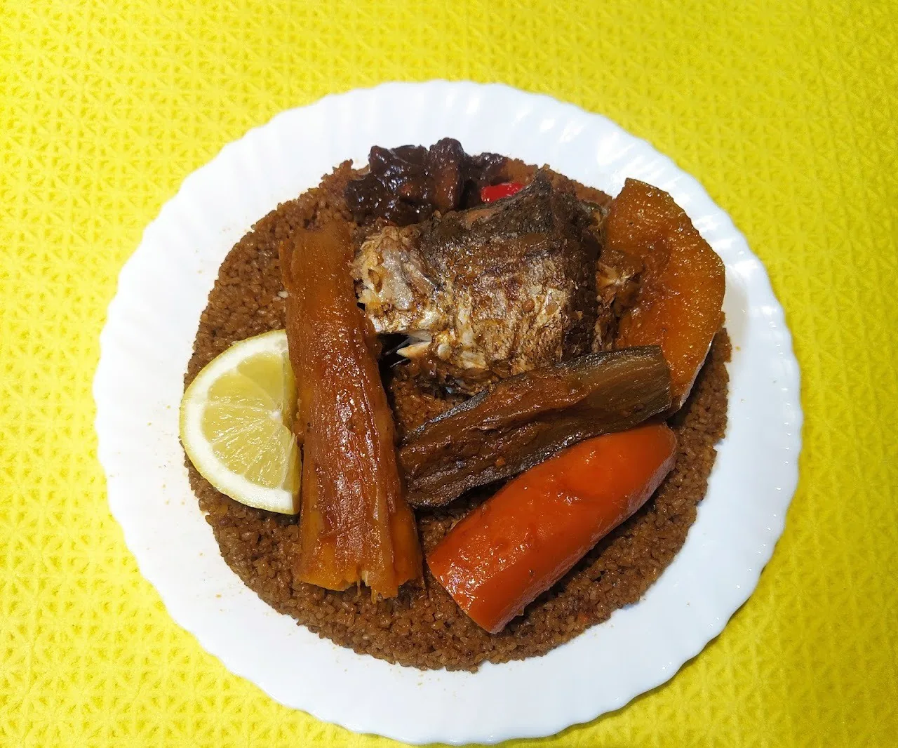 A white plate of grains with a fish piece, carrots and a lemon wedge on a yellow cloth