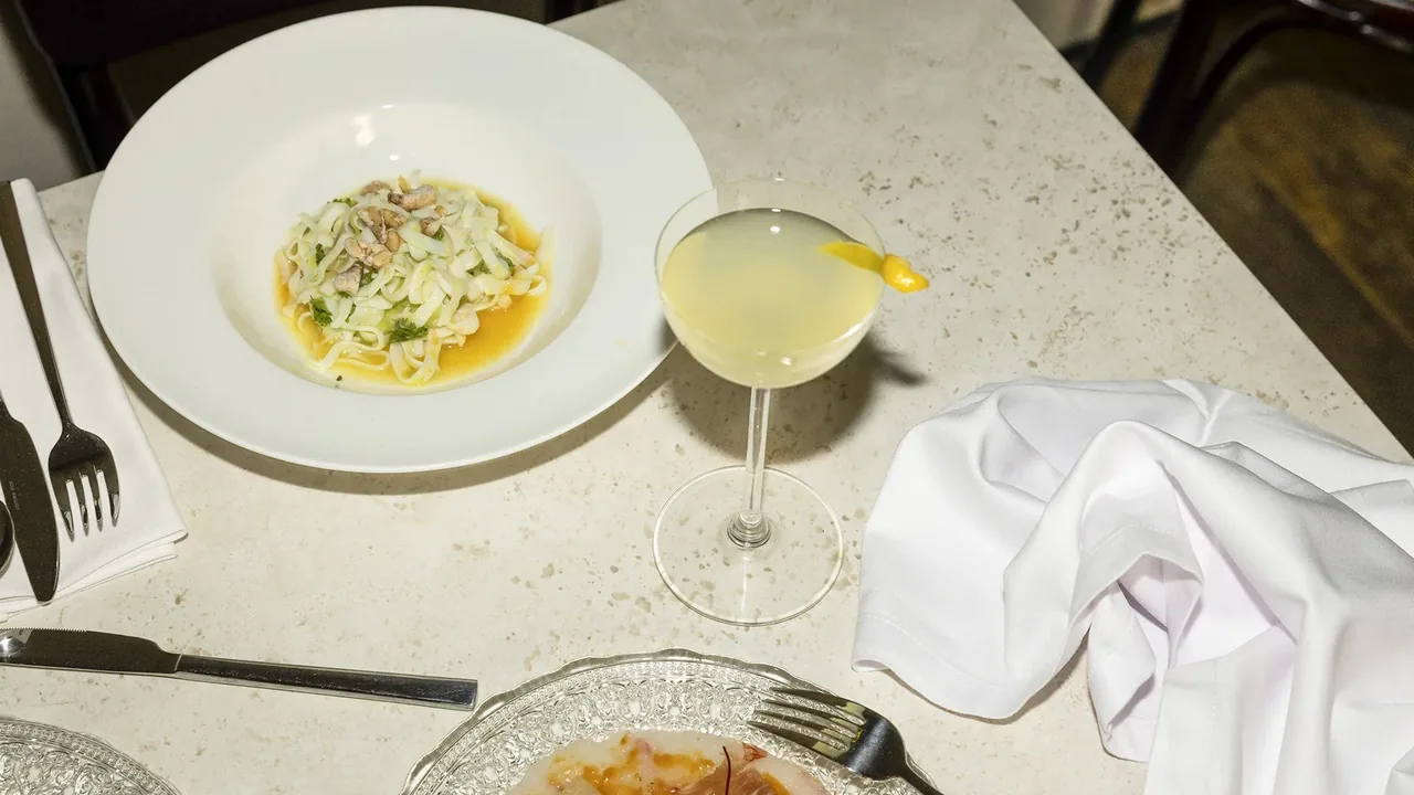 A wide white bowl of fresh ribbon pasta in a light broth with pale shellfish and herbs, beside a coupe of a pale cloudy cocktail with a citrus twist, on a cream terrazzo table with a folded napkin and a silver filigree plate of raw fish below