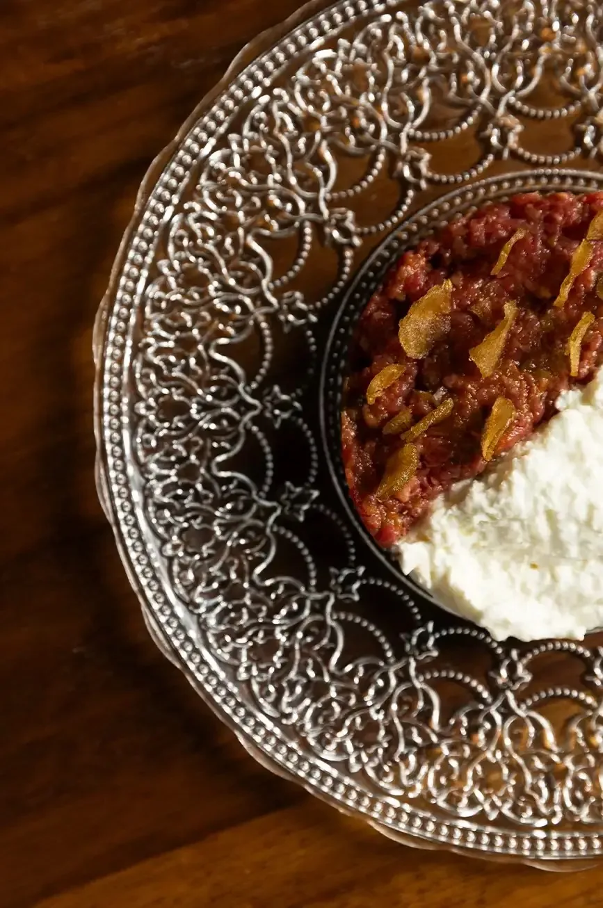 A mound of hand-chopped raw red meat scattered with pale amber shavings, with a spoonful of soft white cheese pressed alongside, served in a dark ring on an ornate silver filigree plate on a wooden table
