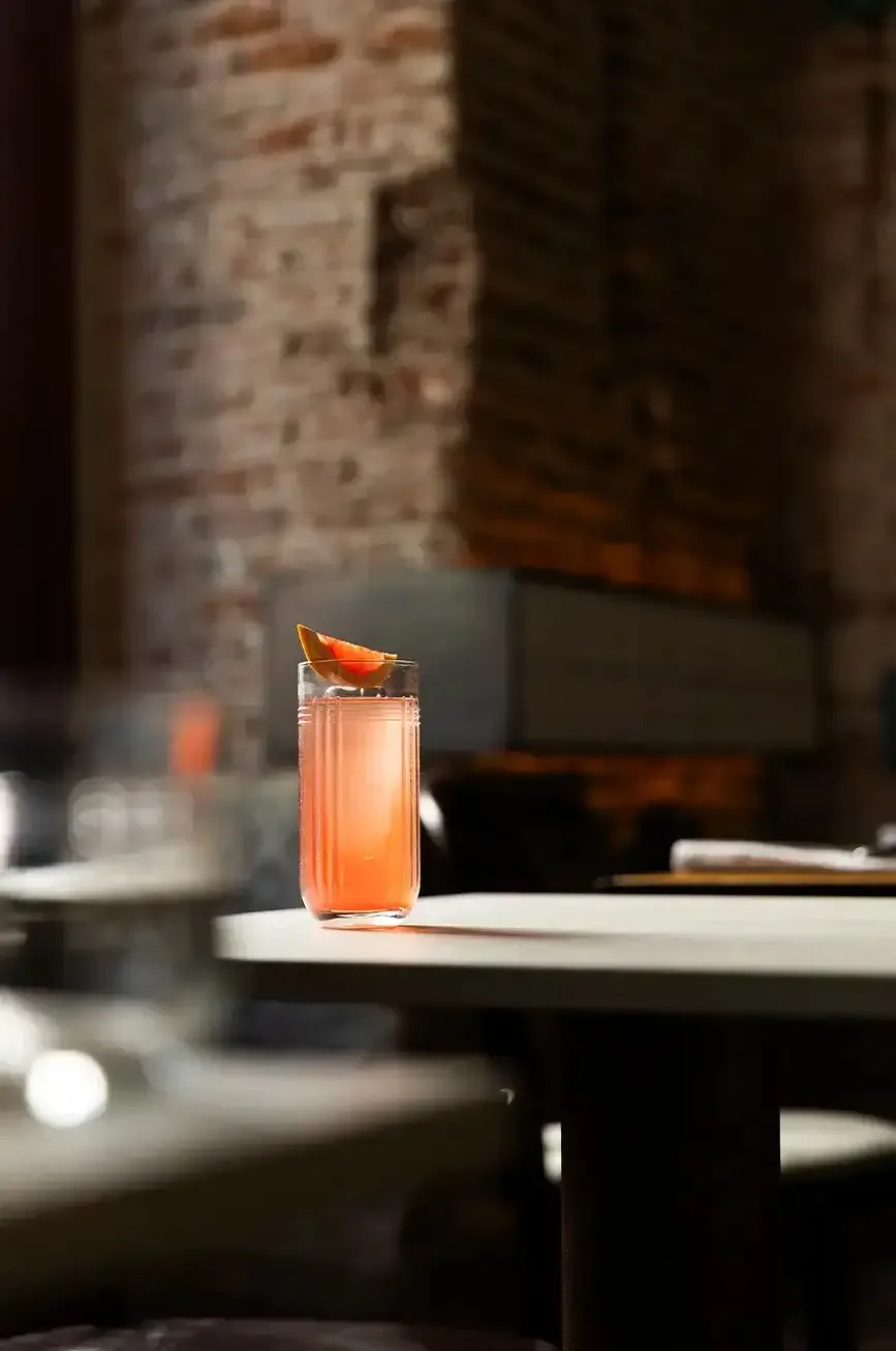 A tall ribbed highball of a pale coral cocktail garnished with a dried citrus wedge, catching a shaft of sunlight on a pale table against an exposed brick wall