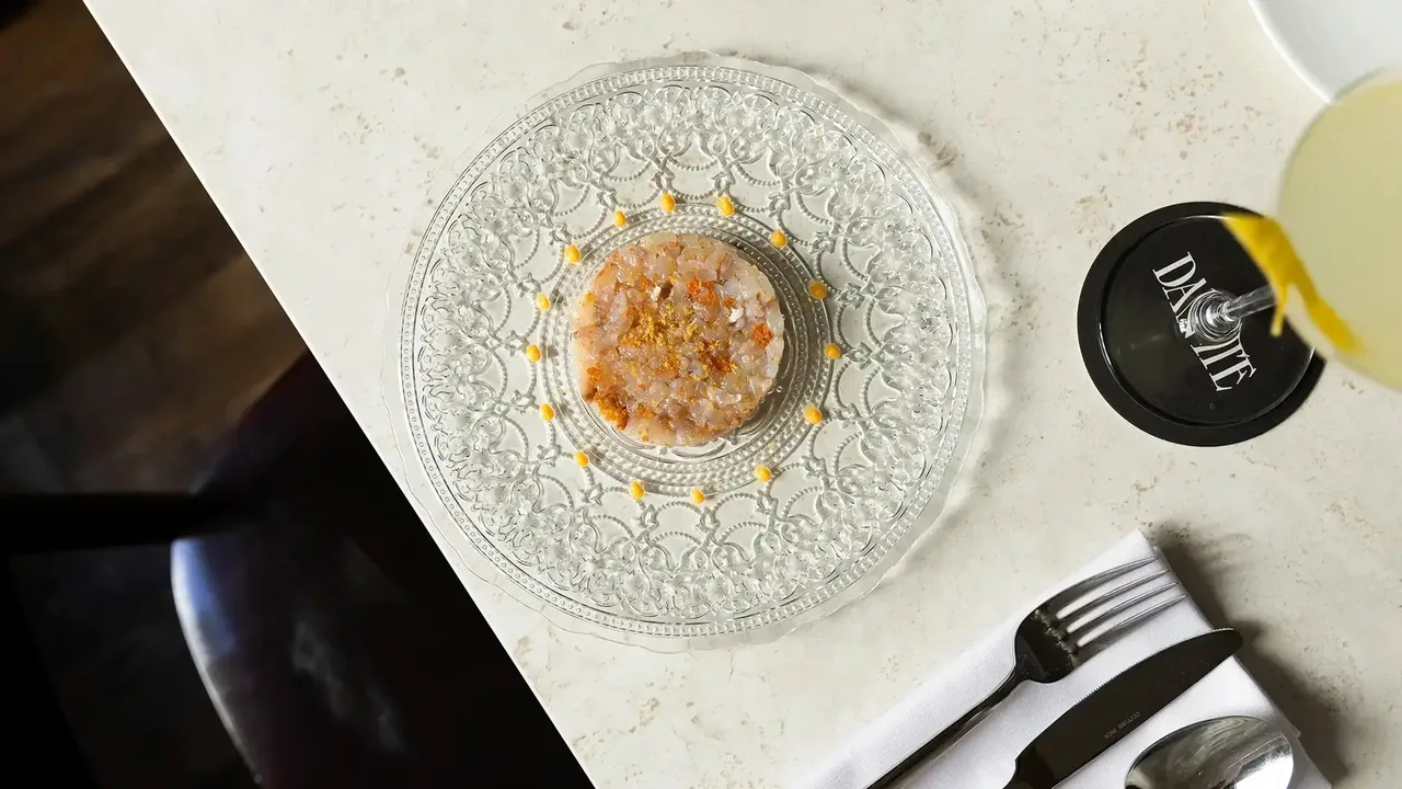 A pressed-glass plate holding a round of finely diced raw fish dusted with orange spice and citrus zest, ringed with dots of yellow sauce, on a cream terrazzo table beside a Dantte-branded coaster under a cocktail glass and a napkin with cutlery