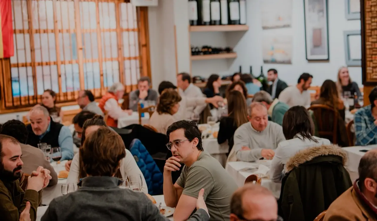 Busy traditional dining room full of seated diners at white-clothed tables with wine racks and framed prints on the walls
