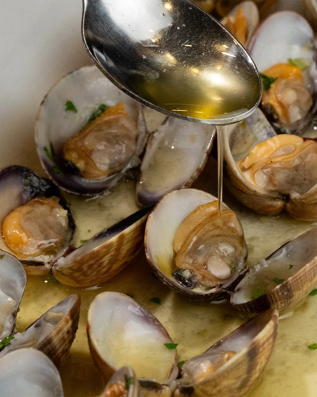 Close-up of clams open in a golden garlic-parsley sauce with a spoon drizzling olive oil over them
