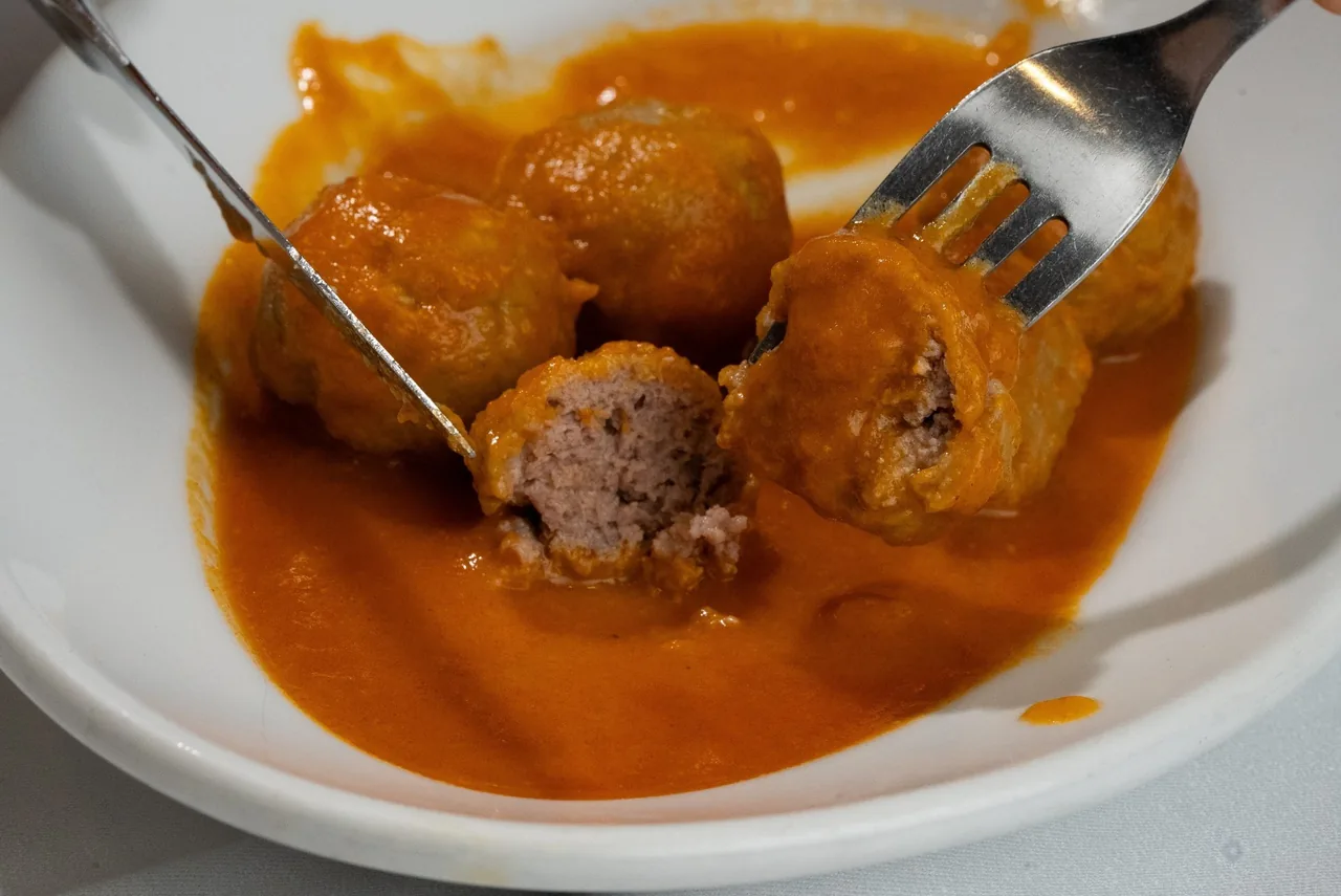 A white plate of meatballs in orange sauce being cut with a fork