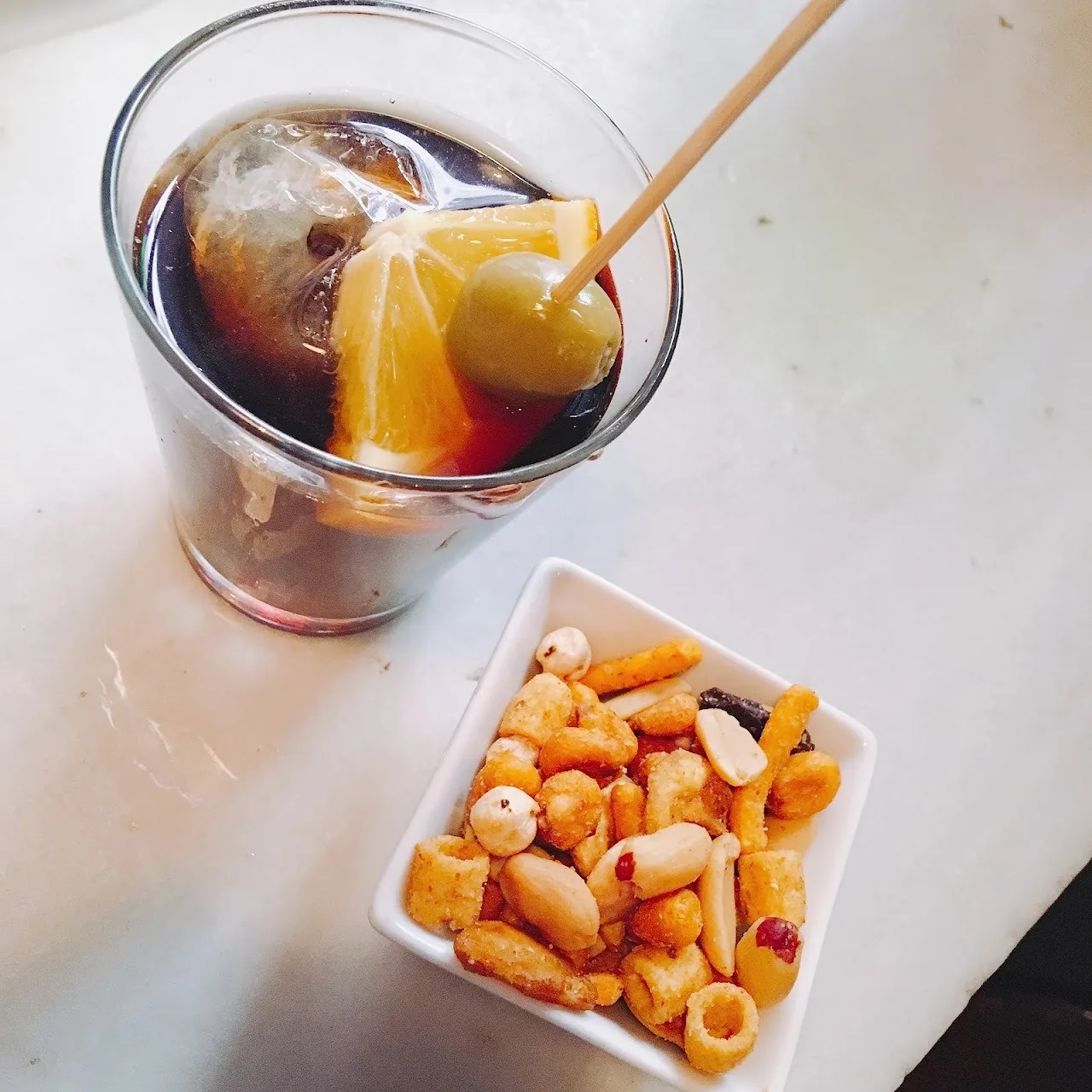 A glass of red vermouth with an orange slice beside a small dish of snacks